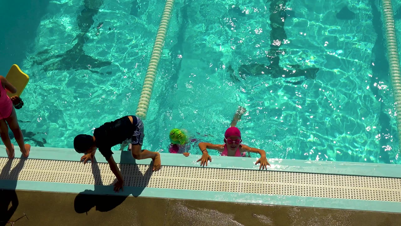 At 0:00 in Cabo San Lucas, Mexico, children in swim caps gather at the pool edge while one child climbs onto the wet deck beside a lane rope.