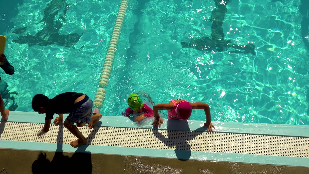 At 0:02 in Cabo San Lucas, Mexico, a child in dark top and patterned shorts moves across the pool deck as two children in bright caps stay at the waterline.