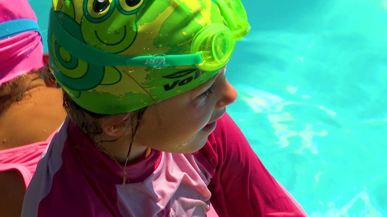 A child in Cabo San Lucas wears a bright green cartoon swim cap and goggles while looking right beside turquoise pool water, with another swimmer partly visible at the left edge.