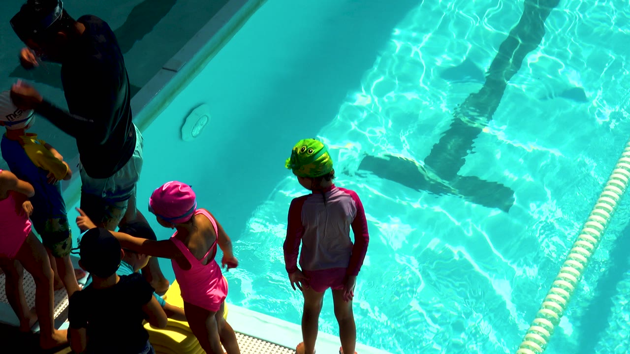 At a pool in Cabo San Lucas, children in colorful swim caps gather along the deck while turquoise water and a lane rope fill the right side.