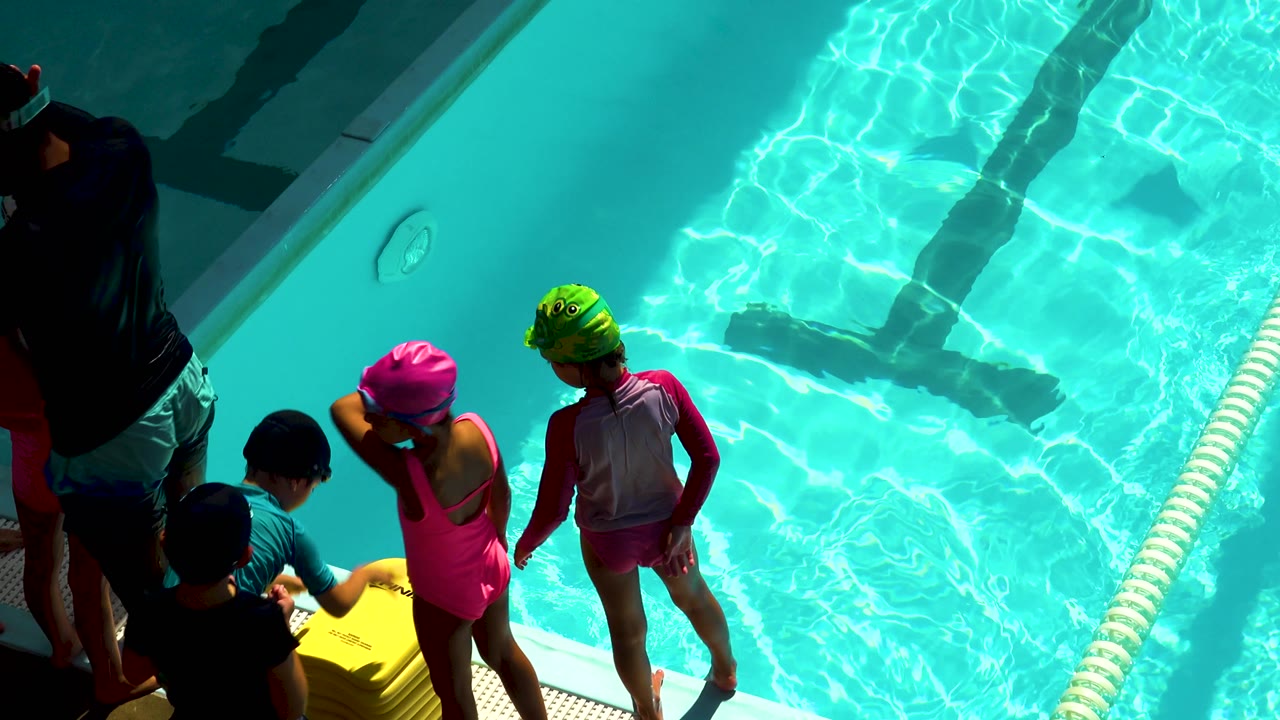 At the Cabo San Lucas pool, a child in a pink swimsuit and pink cap stands beside another child in a green cap while adults and children cluster at the left edge.