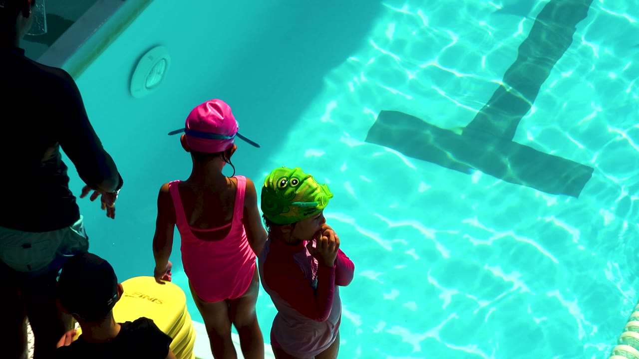 Children wait beside turquoise pool water with an adult nearby.