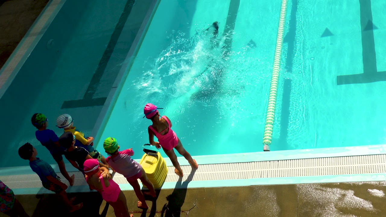 Children in colorful swim caps stand on a wet pool deck in Los Cabos, Mexico, while a burst of water spreads across the turquoise lane pool.