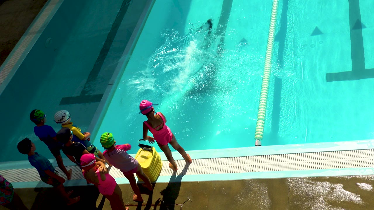 Swim class children watch a splash in a bright outdoor pool.