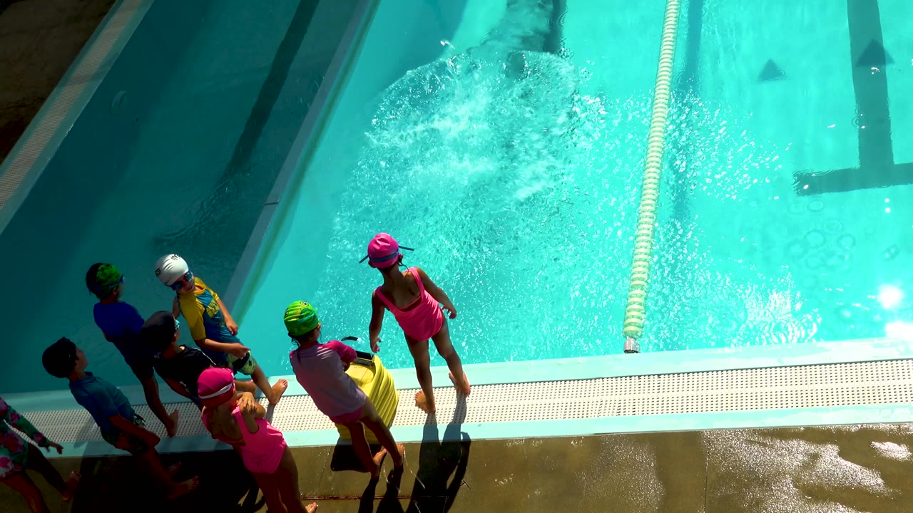 Children in pink, green, and white swim caps stand beside the sunlit Los Cabos pool as foamy water expands across the lane.