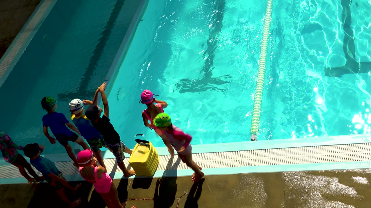 Children in pink, green, white, and dark swim caps stand and move along the deck in Cabo San Lucas, Mexico, while a dark-clad adult is partly visible in the water.