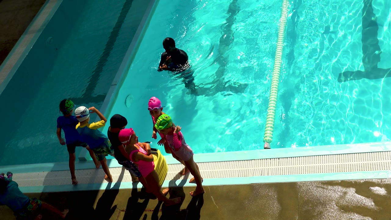 High-angle view in Cabo San Lucas of children in green, pink, white, and yellow swimwear gathered on the wet deck beside turquoise pool water.