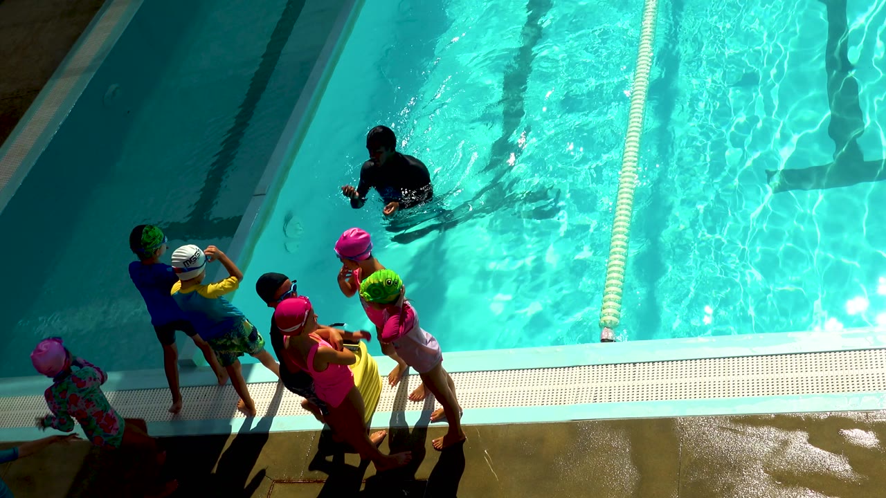 Children in colorful swimwear move along the lower edge of a Cabo San Lucas swimming pool as sunlight ripples across the turquoise lane water.