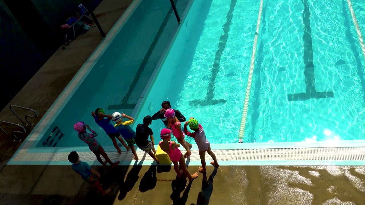 High-angle frame in Cabo San Lucas showing children in colorful swim caps gathered on a wet pool deck beside turquoise lane water.