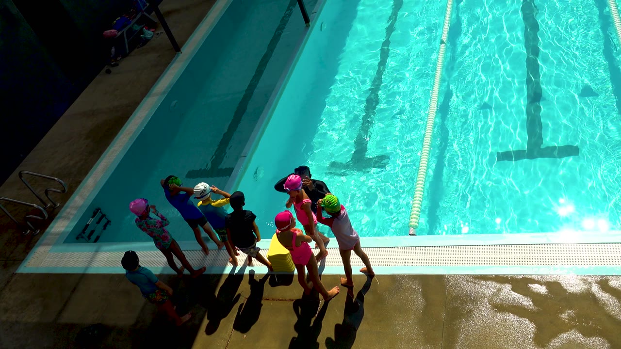 Cabo San Lucas overhead pool view with several children in pink, green, white, and yellow swim caps gathered near the deck drain and lane water.
