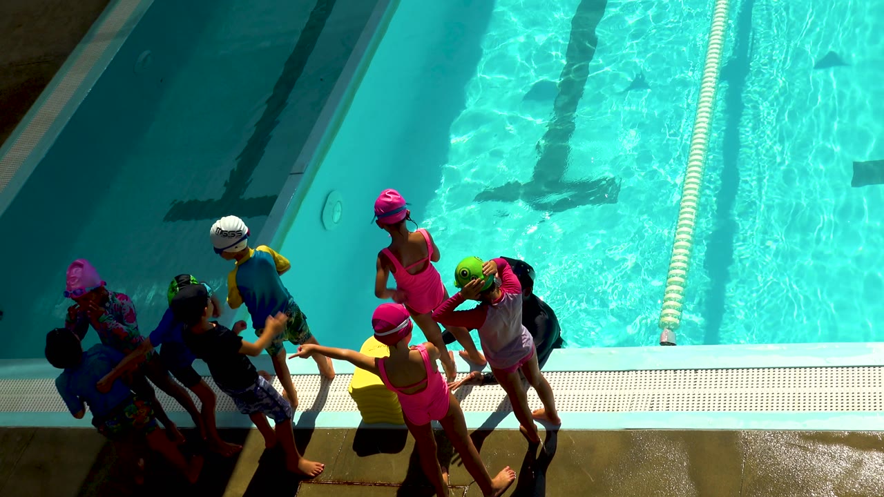 At 0:02 in Cabo San Lucas, Mexico, the group shifts slightly along the pool edge, with a child in a pink swim cap near center and the lane divider running vertically through the water.