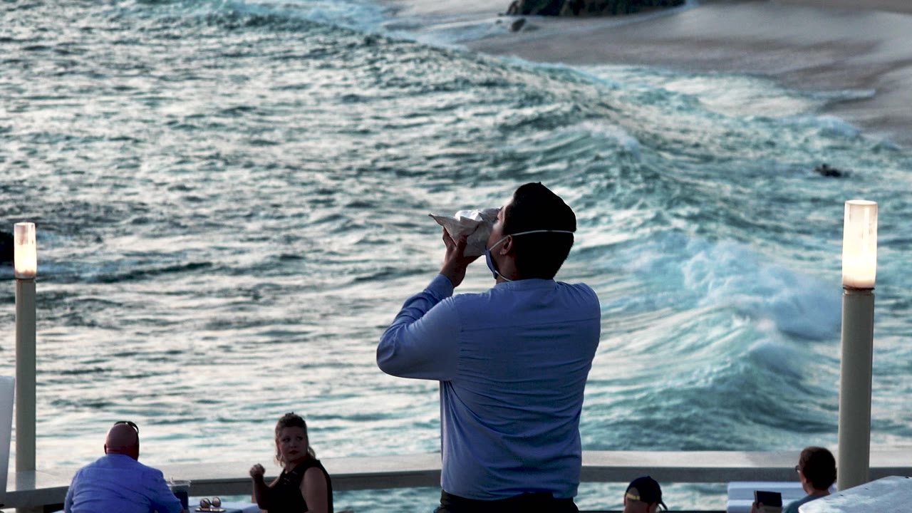 In Cabo San Lucas, Mexico, a man in a blue shirt plays a shell-like mouth instrument near a railing while seated people and foamy surf sit behind him.