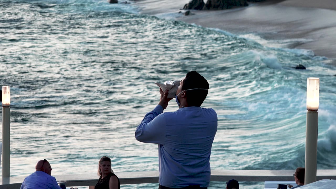 A man holds the shell-like instrument to his mouth near the rail in Cabo San Lucas, Mexico, as a woman looks toward camera and the ocean churns behind them.