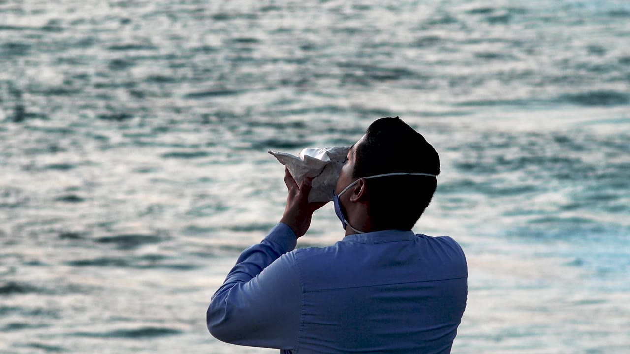 Man seen from behind holding a shell near his face by the sea.