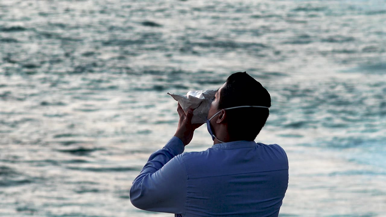 At 0:02, the shell points slightly left as the man stands in the lower right of the Cabo San Lucas frame, backed by darker ripples and pale water highlights.