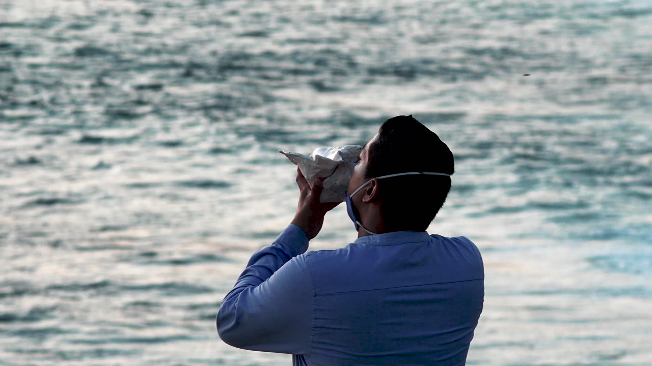 Man in blue shirt holding a conch-like shell in front of ocean water.