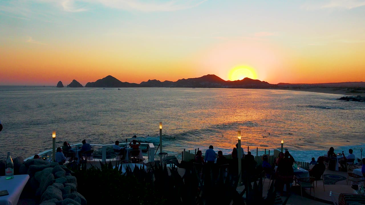 Cabo San Lucas sunset over the ocean with dark coastal rocks on the horizon, terrace lamps and tables in the foreground, and people visible as silhouettes near the railing.