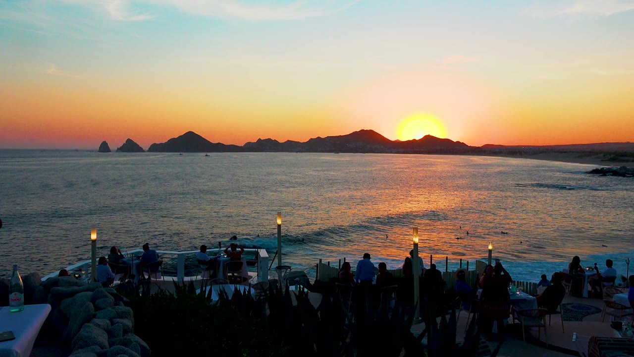 Orange sunset reflecting on the ocean beyond terrace seating and silhouetted people.