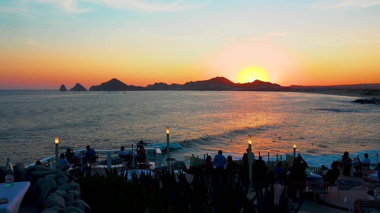 Cabo San Lucas ocean view at sunset with the sun partly behind the dark shoreline, waves rolling toward the lower right, and people seated or standing in silhouette on the terrace.