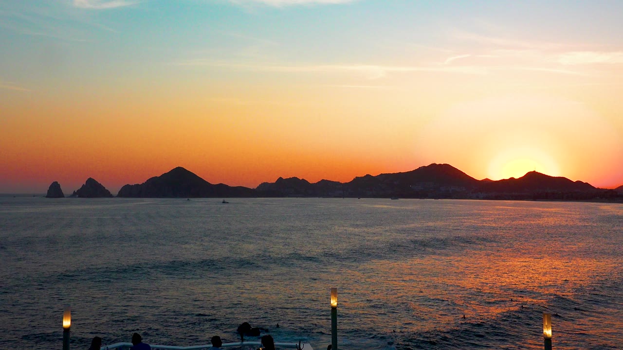 Cabo San Lucas dusk seascape with the sun low behind the mountains, orange reflection spreading across the water, and no visible people.