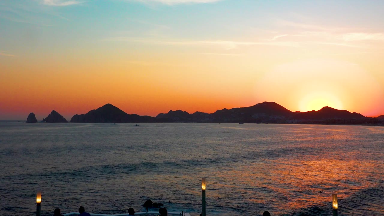 Cabo San Lucas dusk view with a darker ocean foreground, low waves near shore, silhouetted mountains, and no visible people.
