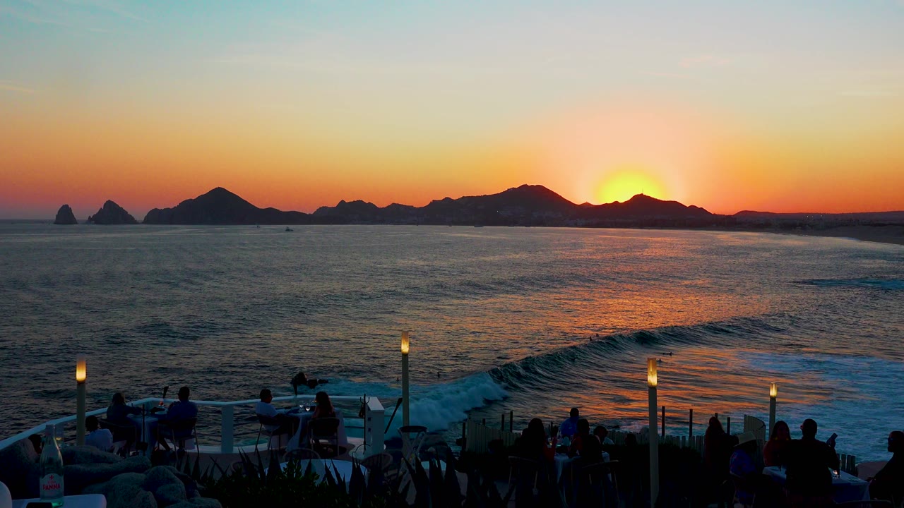 Cabo San Lucas ocean at sunset, with the sun lower against the ridge line and a warm reflection spreading across the darker water.