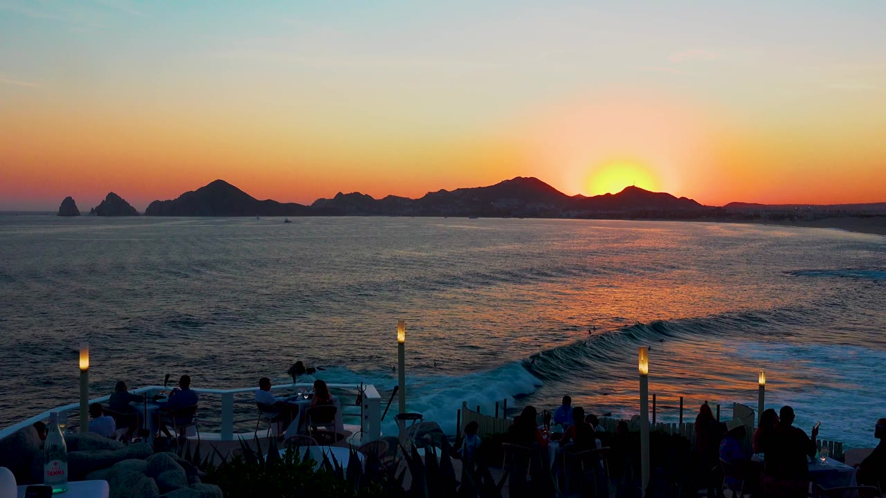 Cabo San Lucas shoreline view with a curling wave in the lower midground, orange sunset light on the water, and silhouetted terrace guests below.