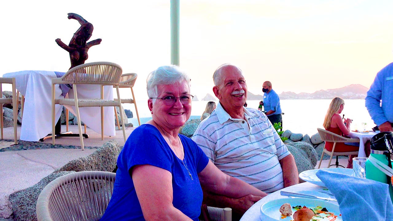Older couple laughing beside a waterfront dining table.