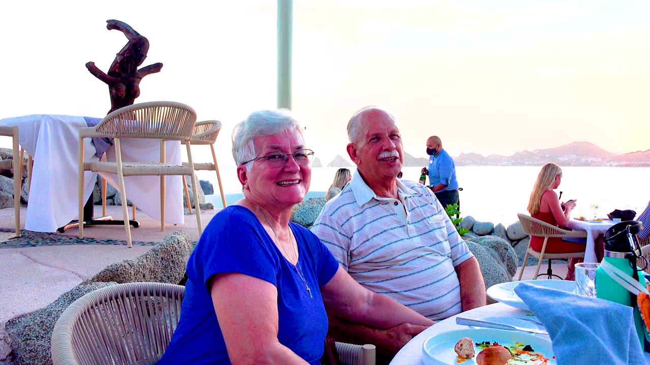 The woman smiles and the man looks outward at a seaside restaurant table in Los Cabos, Mexico, with a seated diner and low hills visible across the water.