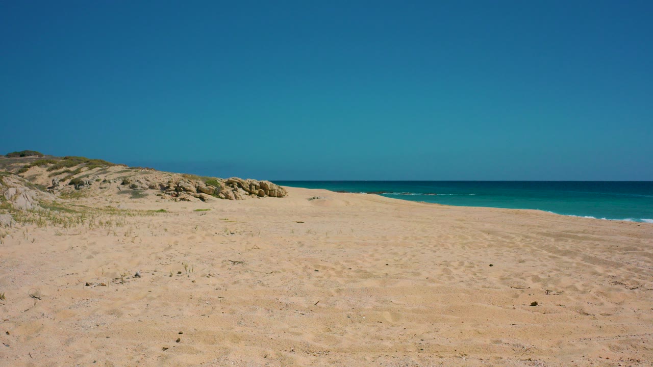 Wide sandy beach with dunes, rocks, blue ocean, and clear sky.