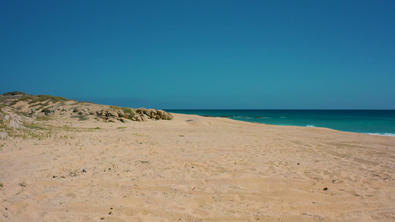 A wide daylight view shows vacant sand at El Cardoncito, a low dune ridge and rocks on the left, blue-green ocean beyond, and no visible people.