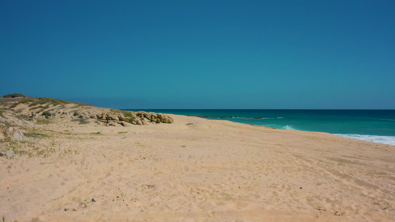 Mid-clip view of El Cardoncito beach with pale sand across the foreground, dunes and rocks along the left edge, turquoise surf at right, and no visible people.