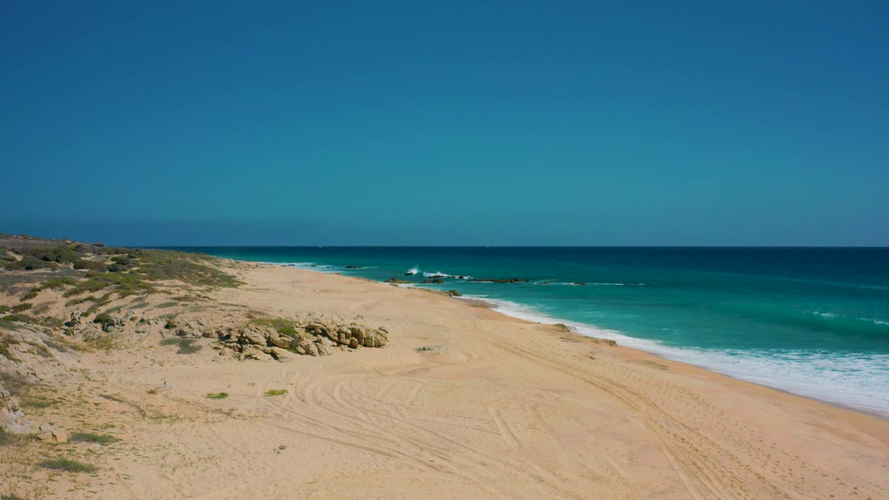 Wide aerial frame over El Cardoncito showing empty sand with curved tire marks, scrub-covered dunes to the left, blue water to the right, and no visible people.