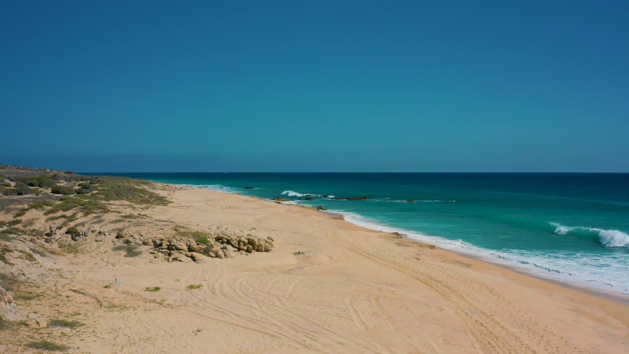 Aerial view of the El Cardoncito shoreline with pale sand, low dunes, turquoise water, a small breaking wave offshore, and no visible people.