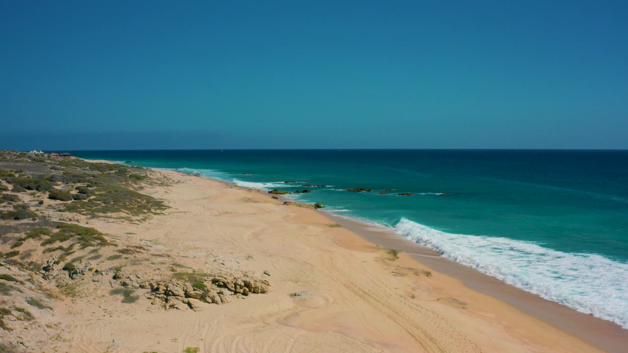 Opening frame of El Cardoncito in Baja California Sur, Mexico, showing pale sand, low scrub, rocks, turquoise water, white surf, and no visible people.