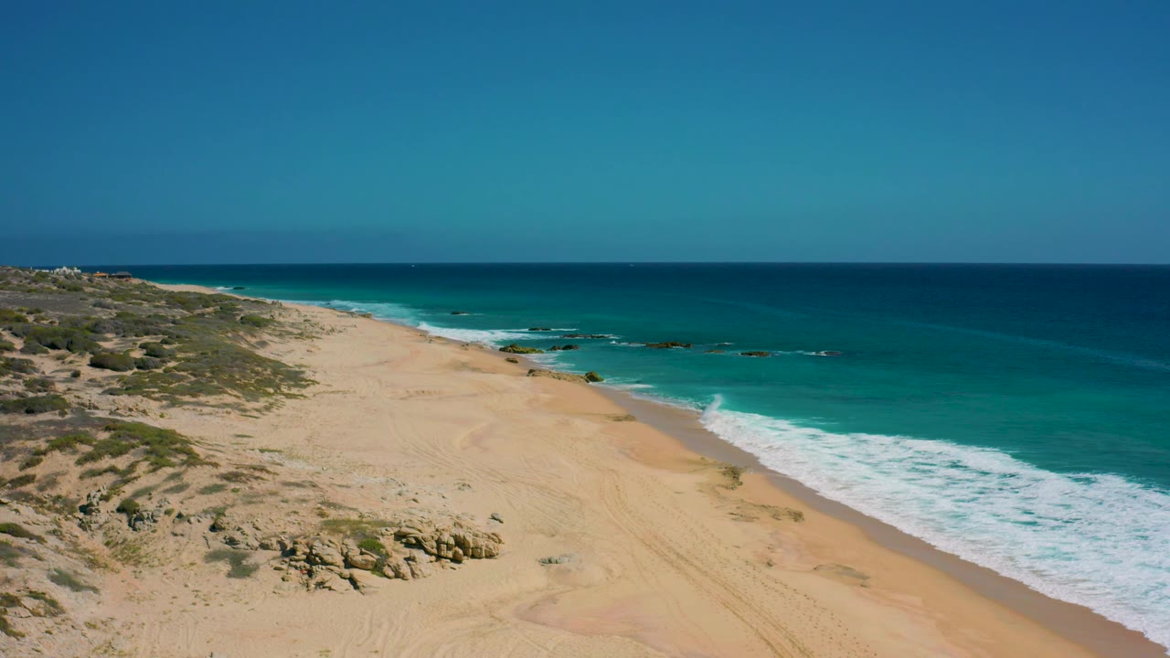 Elevated view over El Cardoncito, Baja California Sur, Mexico, where pale sand and rocky patches meet turquoise shallows and a band of white surf, with no visible people.