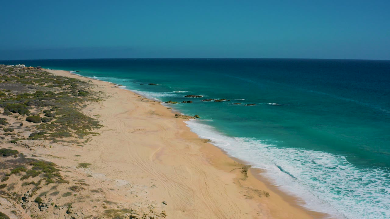Opening aerial frame over El Cardoncito beach in Baja California Sur, with pale sand, scrub-covered dunes, turquoise surf, darker offshore water, and no visible people.