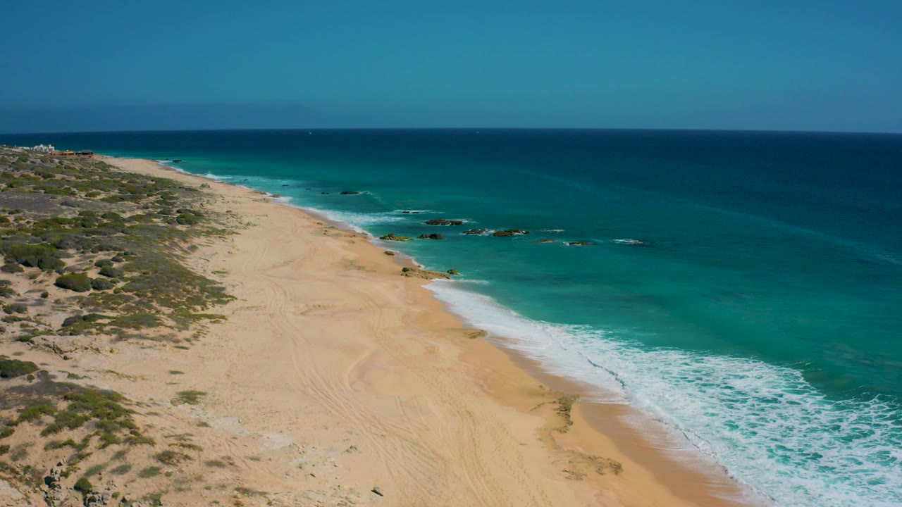Aerial view of El Cardoncito beach with the surf band pushed farther along the right side, empty tan sand in the foreground, scrubland to the left, and no visible people.