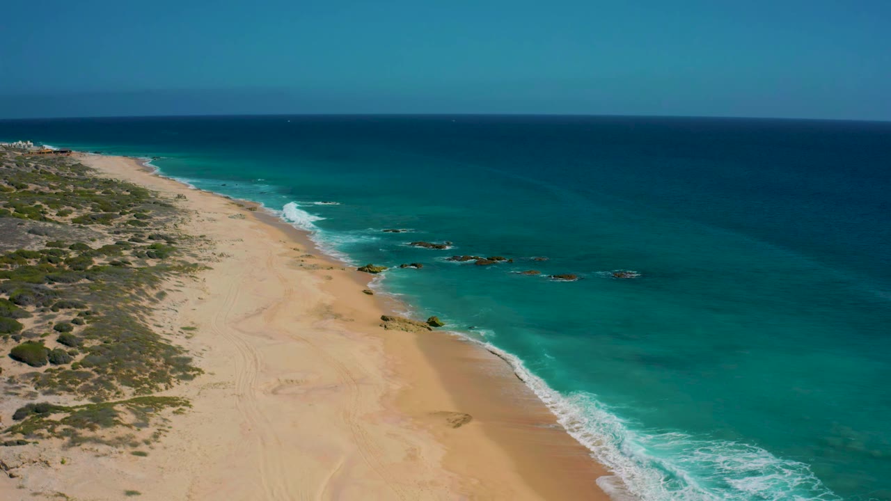 Aerial view at El Cardoncito showing the shoreline angled through frame, pale sand beside clear green-blue water, small rocky outcrops offshore, and no visible people.