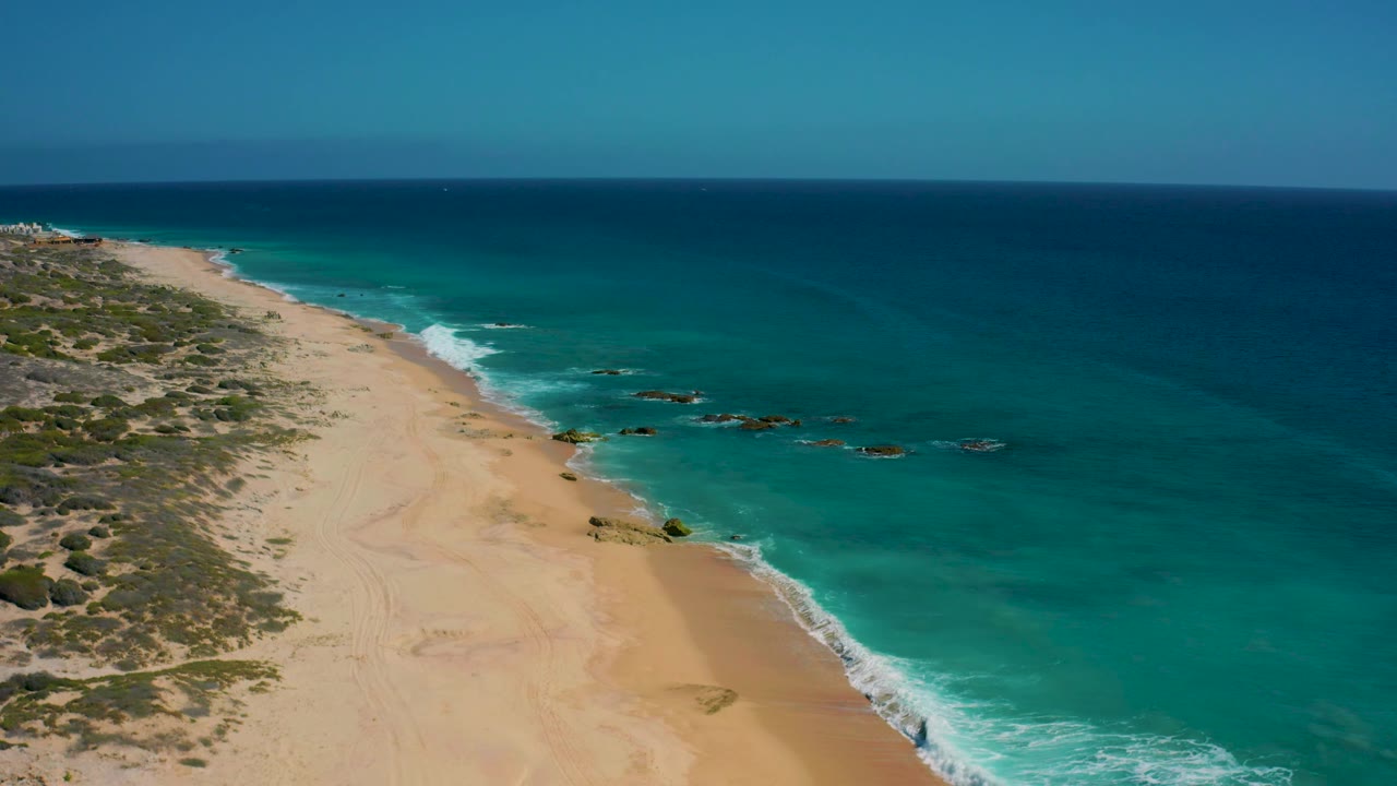 Aerial frame from El Cardoncito with empty sand curving along the left edge, bright turquoise shallows around offshore rocks, deeper blue sea beyond, and no visible people.