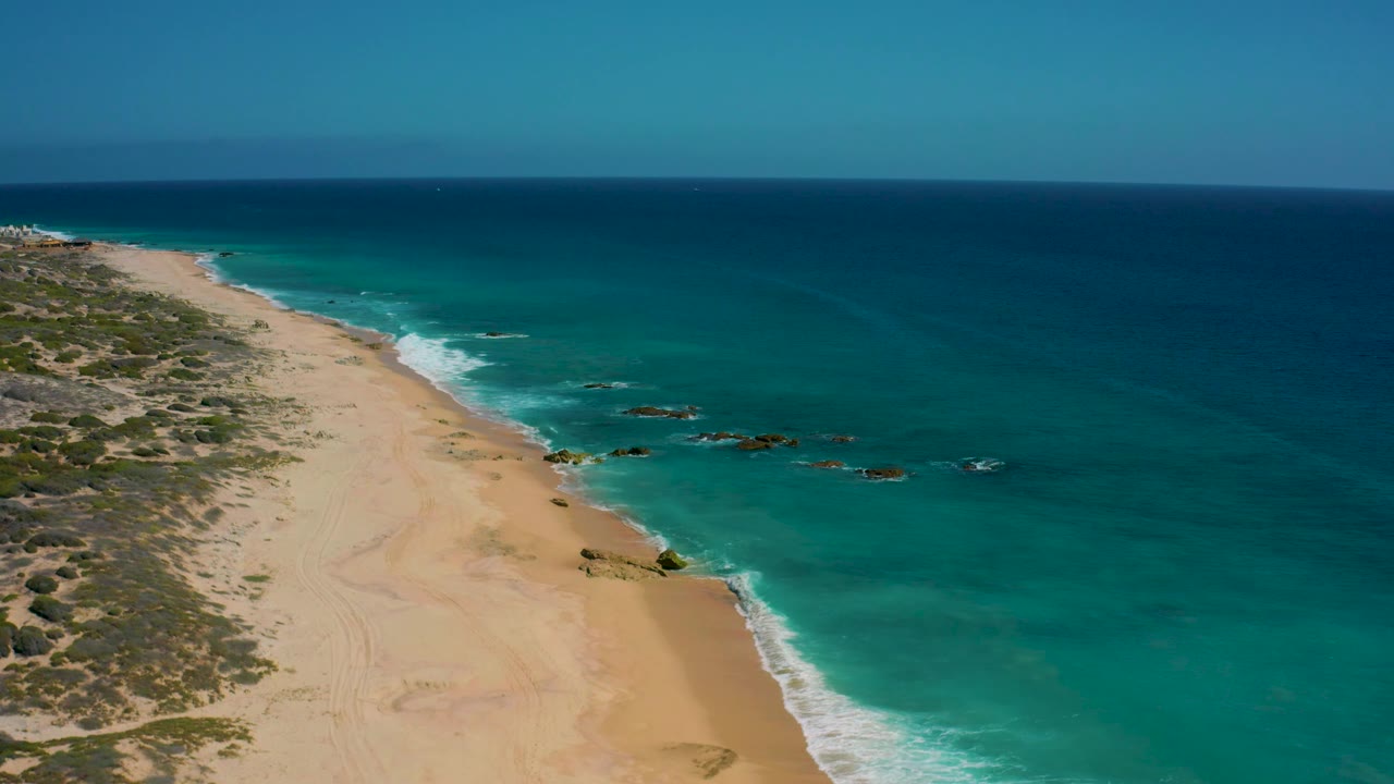 Aerial view over El Cardoncito with scrub-covered land and sand on the left, a narrow white surf line, rocky turquoise shallows in the center, open ocean to the right, and no visible people.