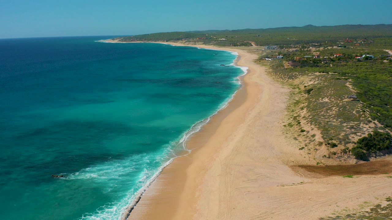 Aerial frame over El Cardoncito, Baja California Sur, with blue-green water on the left, a broad tan beach in the center, low dunes and vegetation on the right, and no visible people.