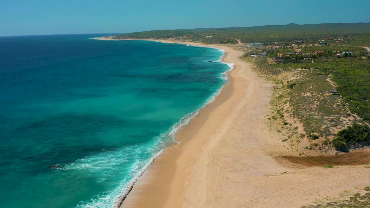 Blue ocean waves curve along a tan beach bordered by scrub vegetation.