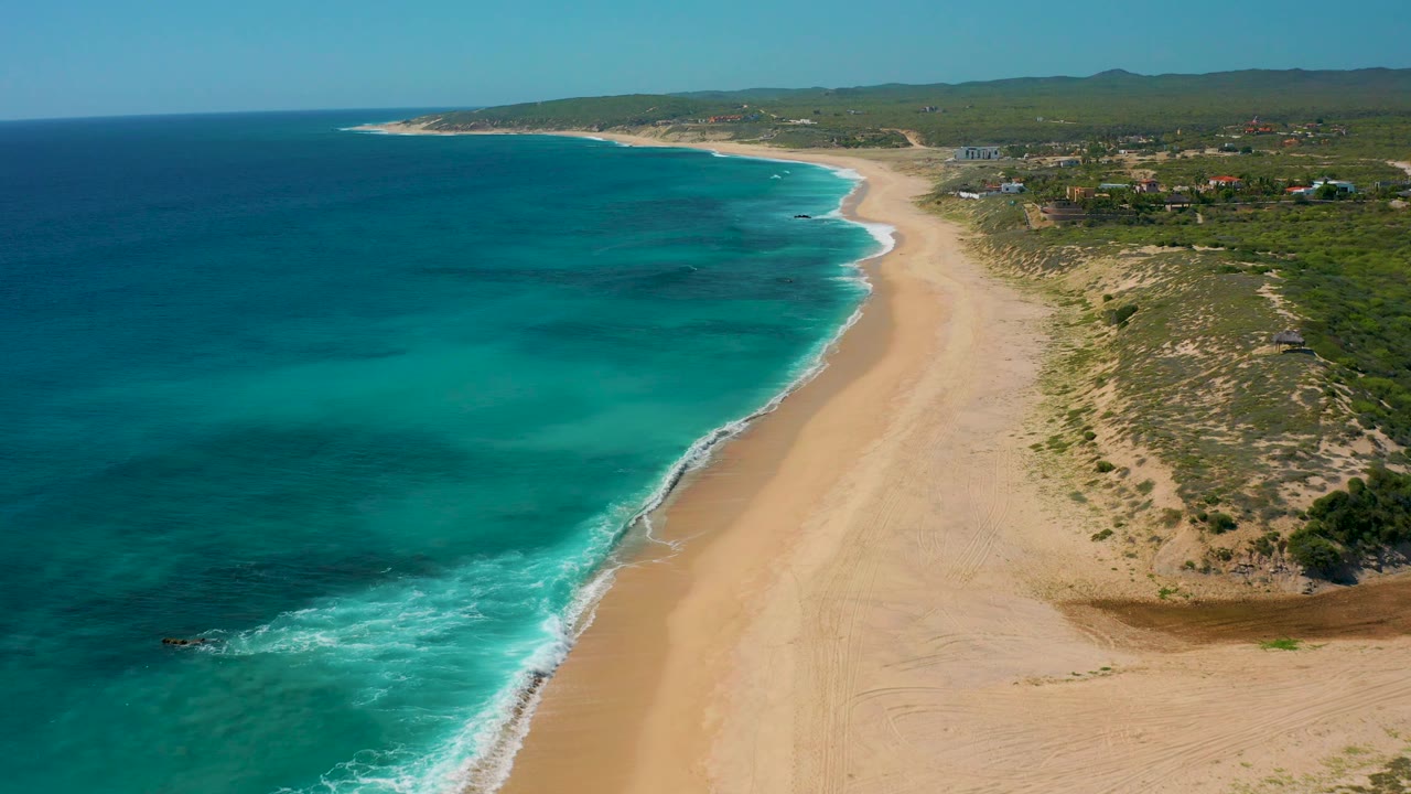 The El Cardoncito beach fills the frame with turquoise water meeting tan sand, dune vegetation to the right, scattered inland buildings near the horizon, and no visible people.