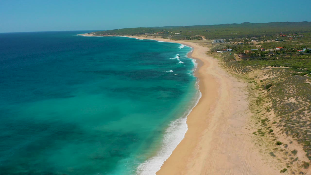 Aerial daylight view at El Cardoncito, Baja California Sur, with turquoise water on the left, a broad sandy beach, scrub dunes, distant buildings, low hills, and no visible people.
