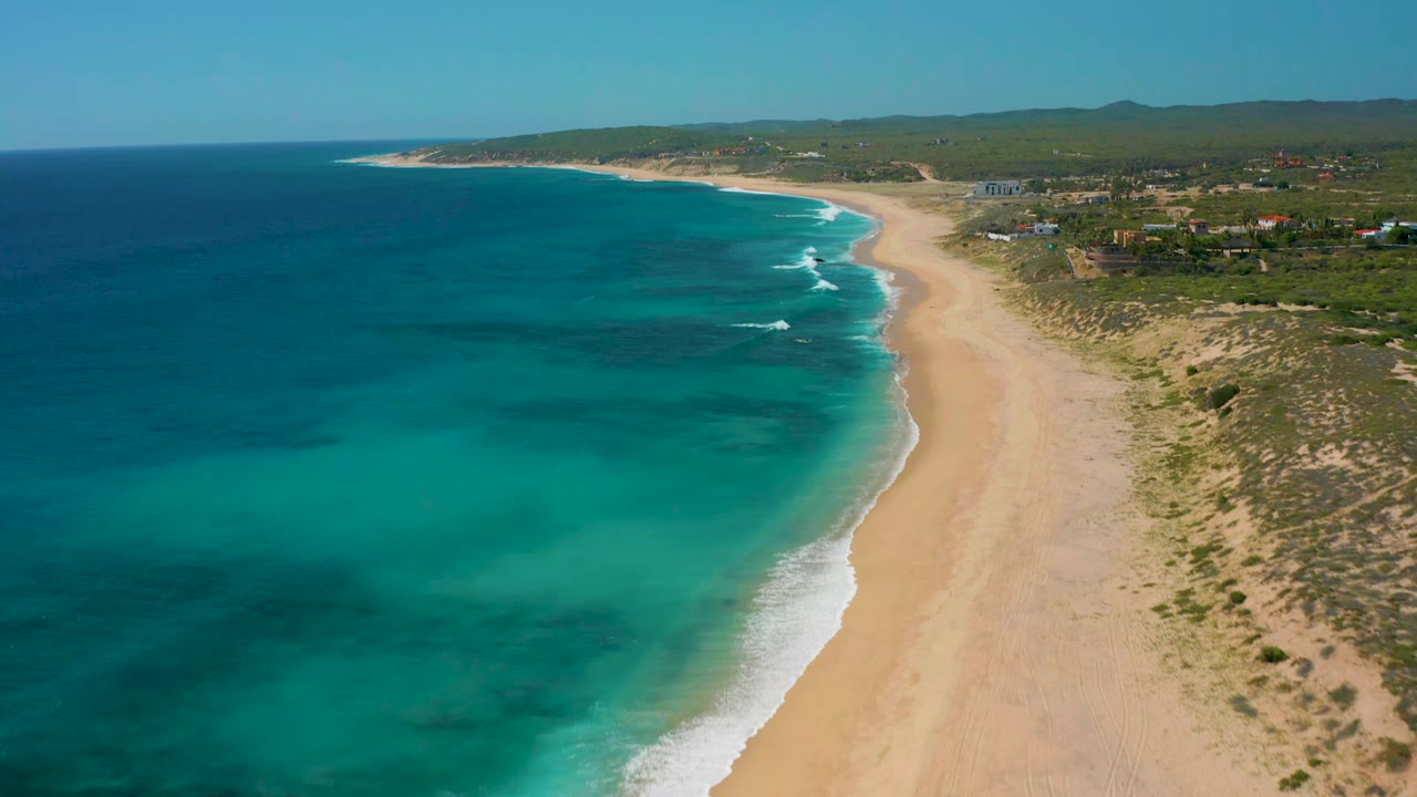 Drone view of clear blue water and surf meeting a long sandy shoreline.