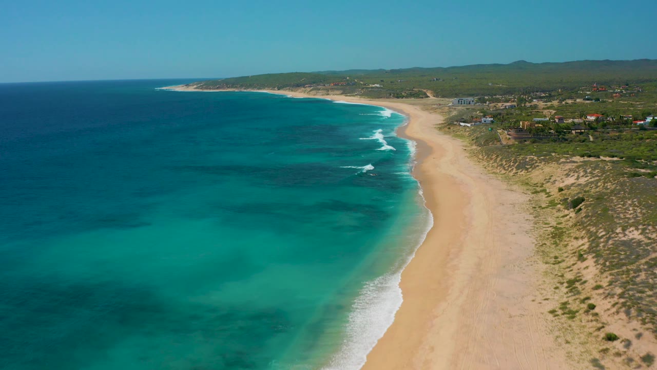 Aerial frame over El Cardoncito with darker offshore water patches, bright turquoise shallows, a pale sandy beach, scrub-covered dunes, scattered inland buildings, and no visible people.
