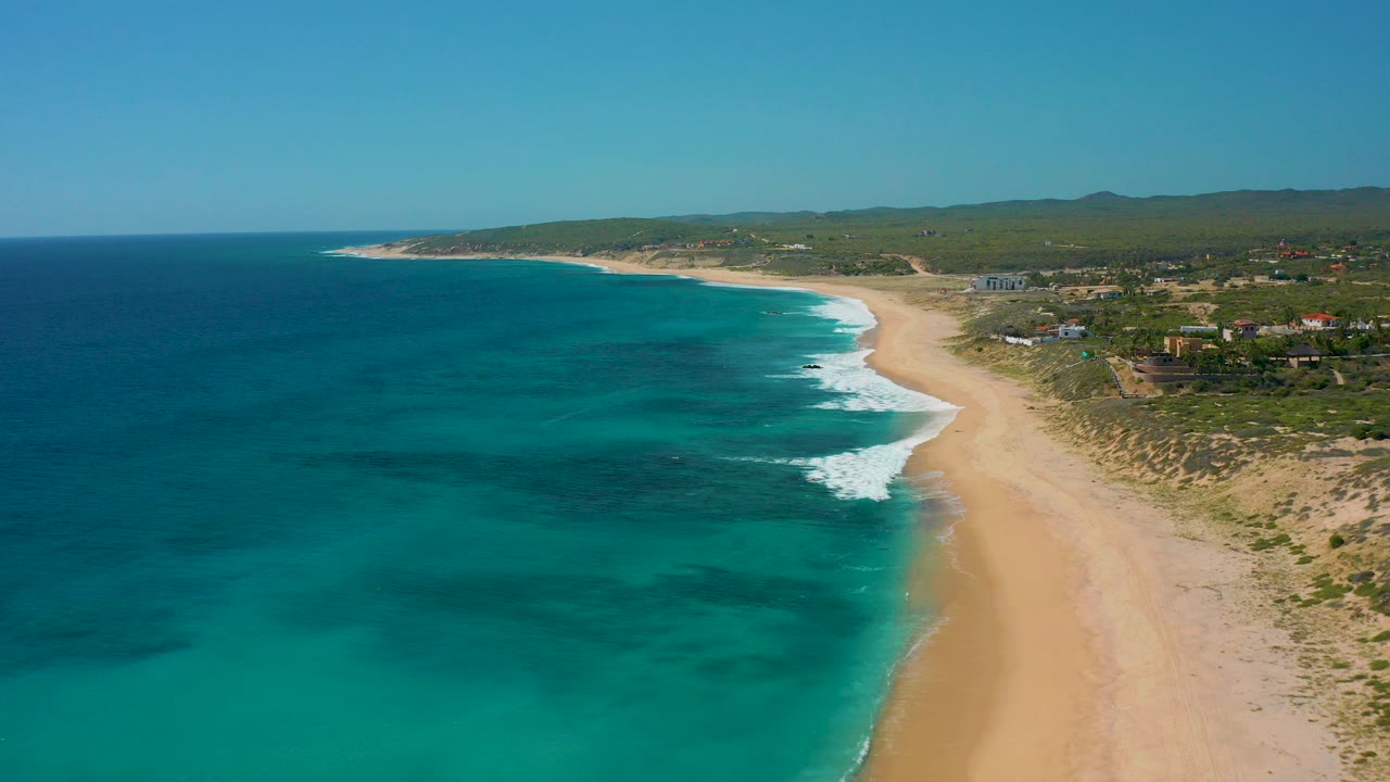 Aerial view over El Cardoncito, Baja California Sur, showing deep blue water, a pale sandy beach, low greenery, scattered houses, and no visible people at the start of the clip.