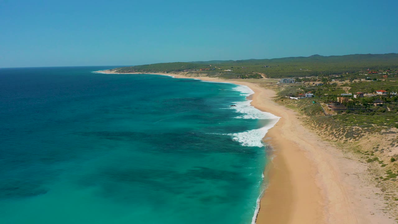 High aerial frame over El Cardoncito with dark patches in blue-green water, white surf touching the sand, low vegetation, small coastal buildings, and no visible people.