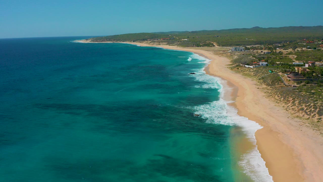 Aerial frame over El Cardoncito, Baja California Sur, showing blue-green water, a pale sandy shoreline, low coastal vegetation, distant buildings, and no visible people.